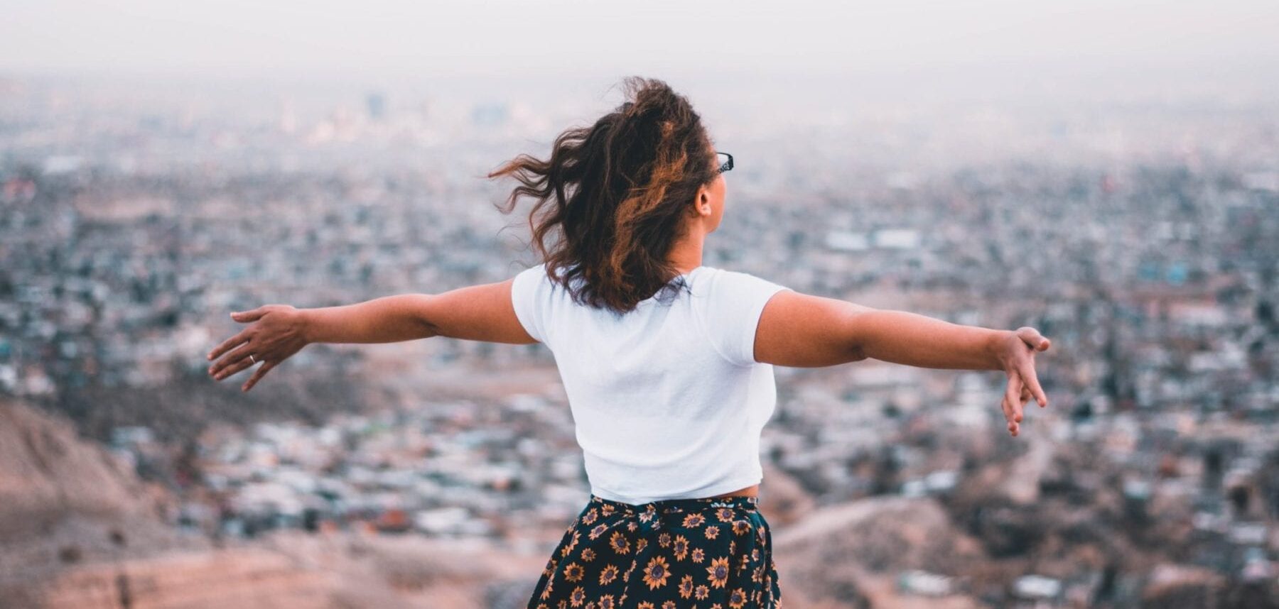 Woman on hill overlooking city below. She is standing with arms open wide, feeling free.